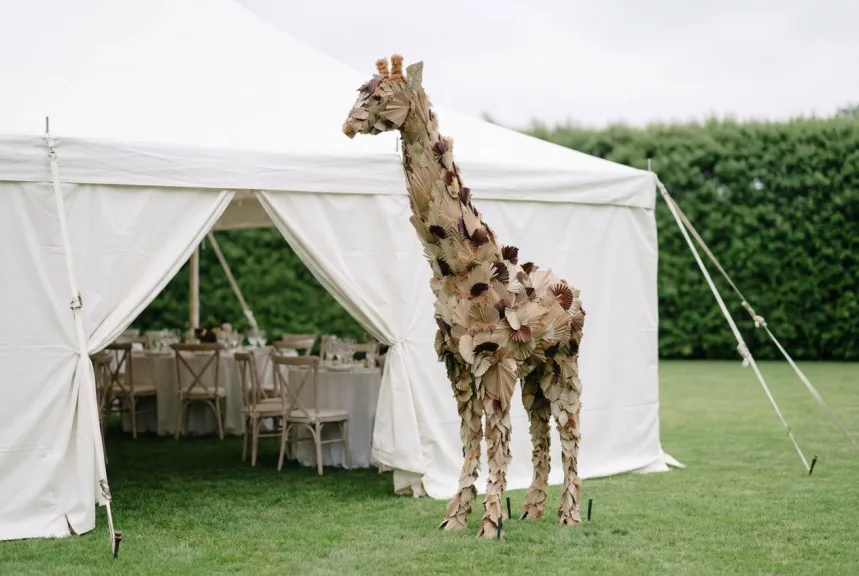 A very tall giraffe sculpture covered in dried palm leaves beside a white event tent