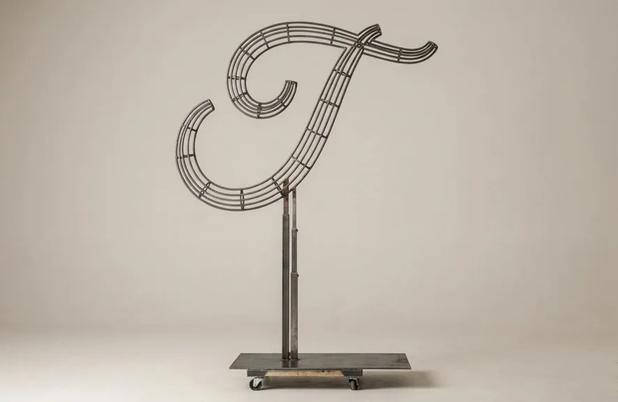 Monogram letter T topiary frame in Flowing script style, bare galvanized steel on an adjustable stand