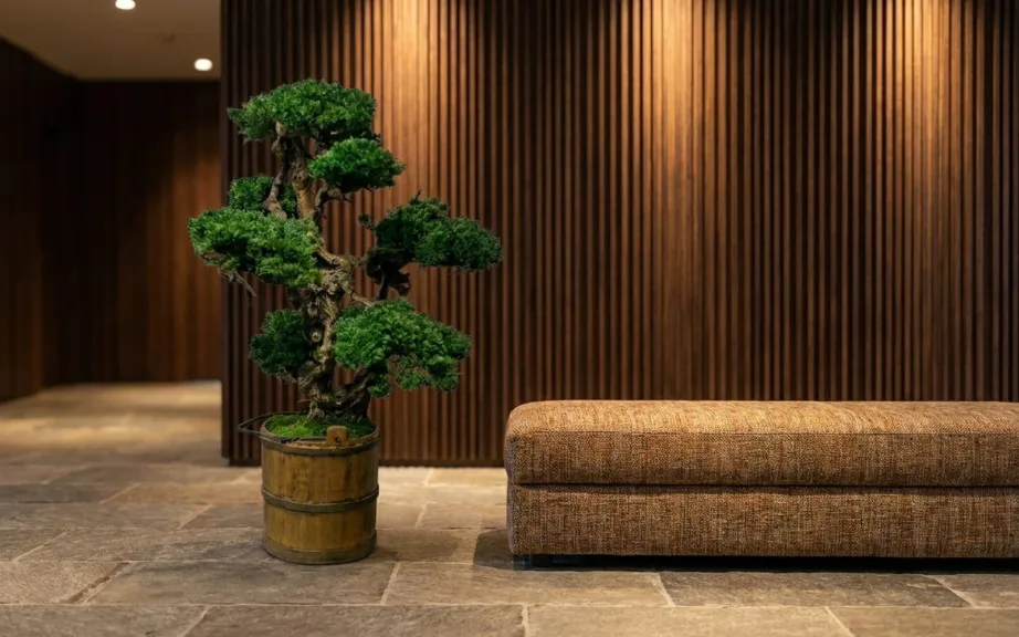 A tall preserved bonsai with several foliage pads in a wooden tub on the stone floor of a hotel lobby, beside an upholstered bench.